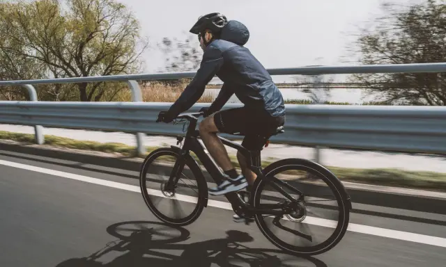 Un cycliste sur un vélo électrique qui se recharge en roulant, longeant une voie rapide bordée d'eau et d'arbres.