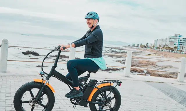 Une femme souriante roule sur un vélo électrique, profitant de la vue sur la mer. Le poid du vélo électrique ne l'empêche pas de profiter de sa balade.