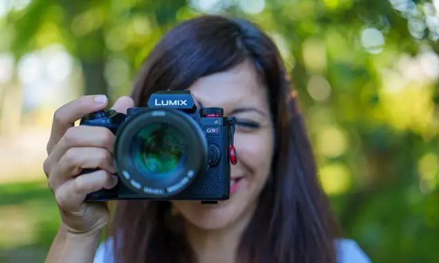 Une femme souriante tient un appareil photo Lumix G9II devant son visage. Un guide pour comment choisir son appareil photo.