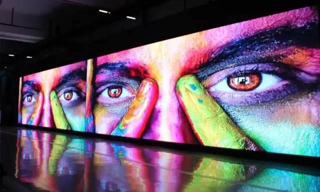Un écran géant affiche un visage aux yeux intenses, couvert de peinture colorée. La dalle IPS ou LED rend les couleurs vibrantes et le reflet sur le sol ajoute à l'effet visuel.