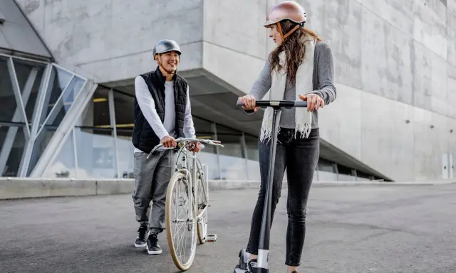 Un homme à vélo et une femme en trottinette, tous deux portant un casque obligatoire, discutent devant un bâtiment moderne.