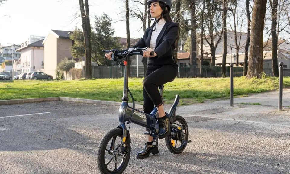 Femme en costume, casque, assise sur une trottinette Urban Glide qui n'avance plus. Elle semble dépitée.