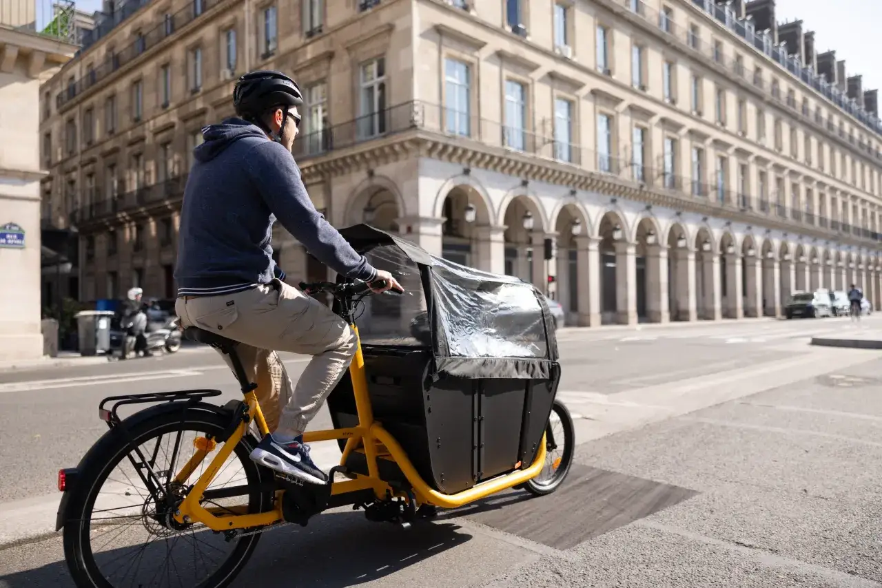 Un homme roule à Paris sur un vélo cargo électrique jaune, son poids est bien réparti pour une conduite stable.