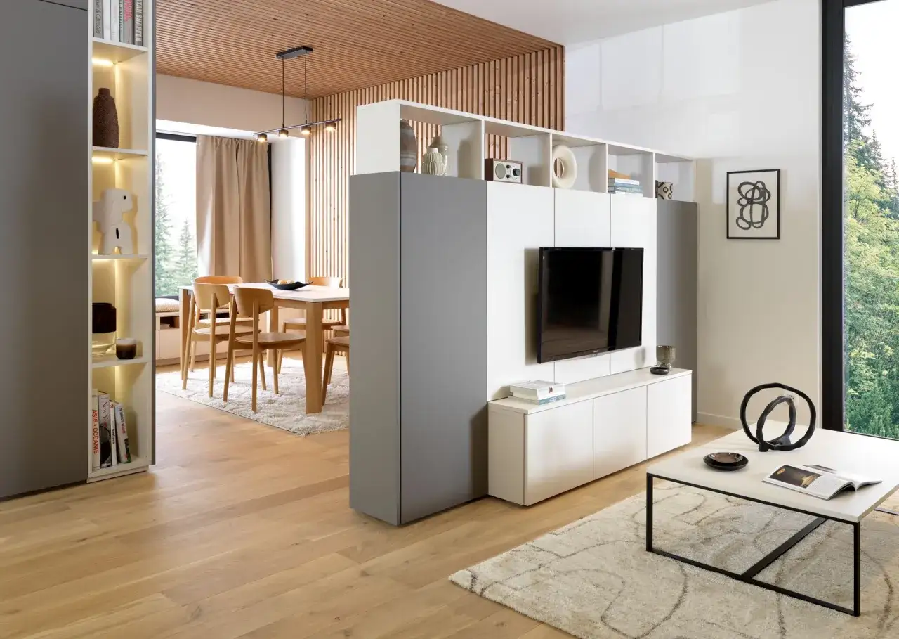 Salon moderne avec une smart TV intégrée dans une étagère murale. Table basse, salle à manger et vue sur la forêt.