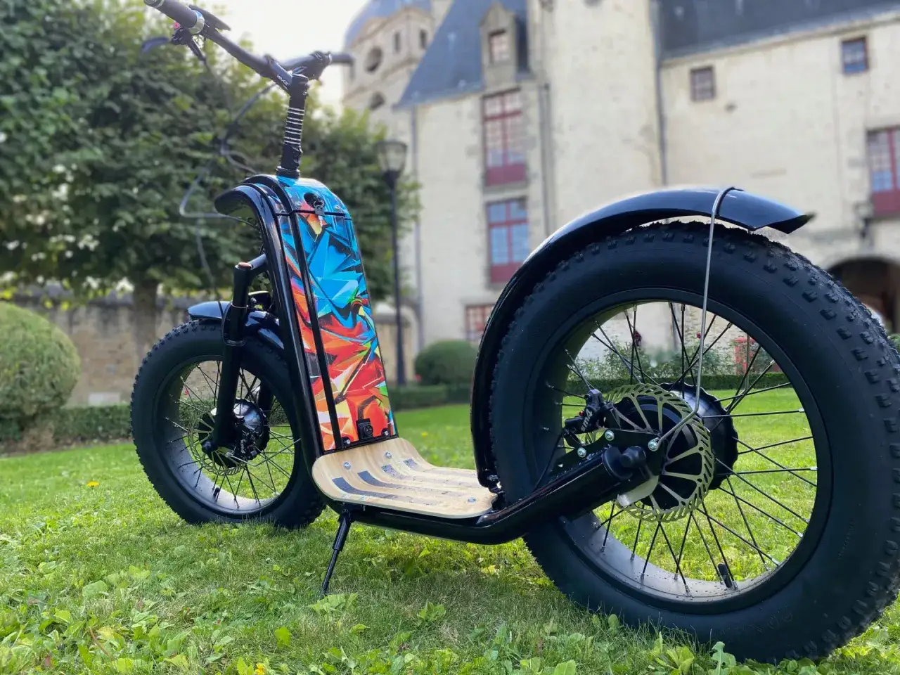 Trottinette manuelle noire avec un plateau en bois et un design coloré sur le garde-boue, sur fond d'herbe et de bâtiment.